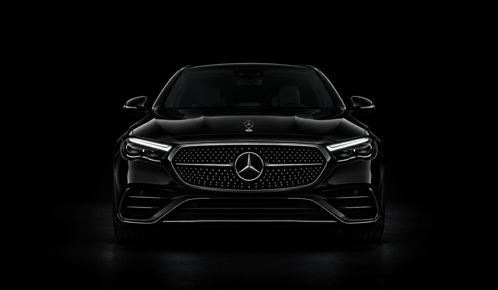 Black Mercedes-Benz E-Class saloon, front view, headlights on, against a black studio background