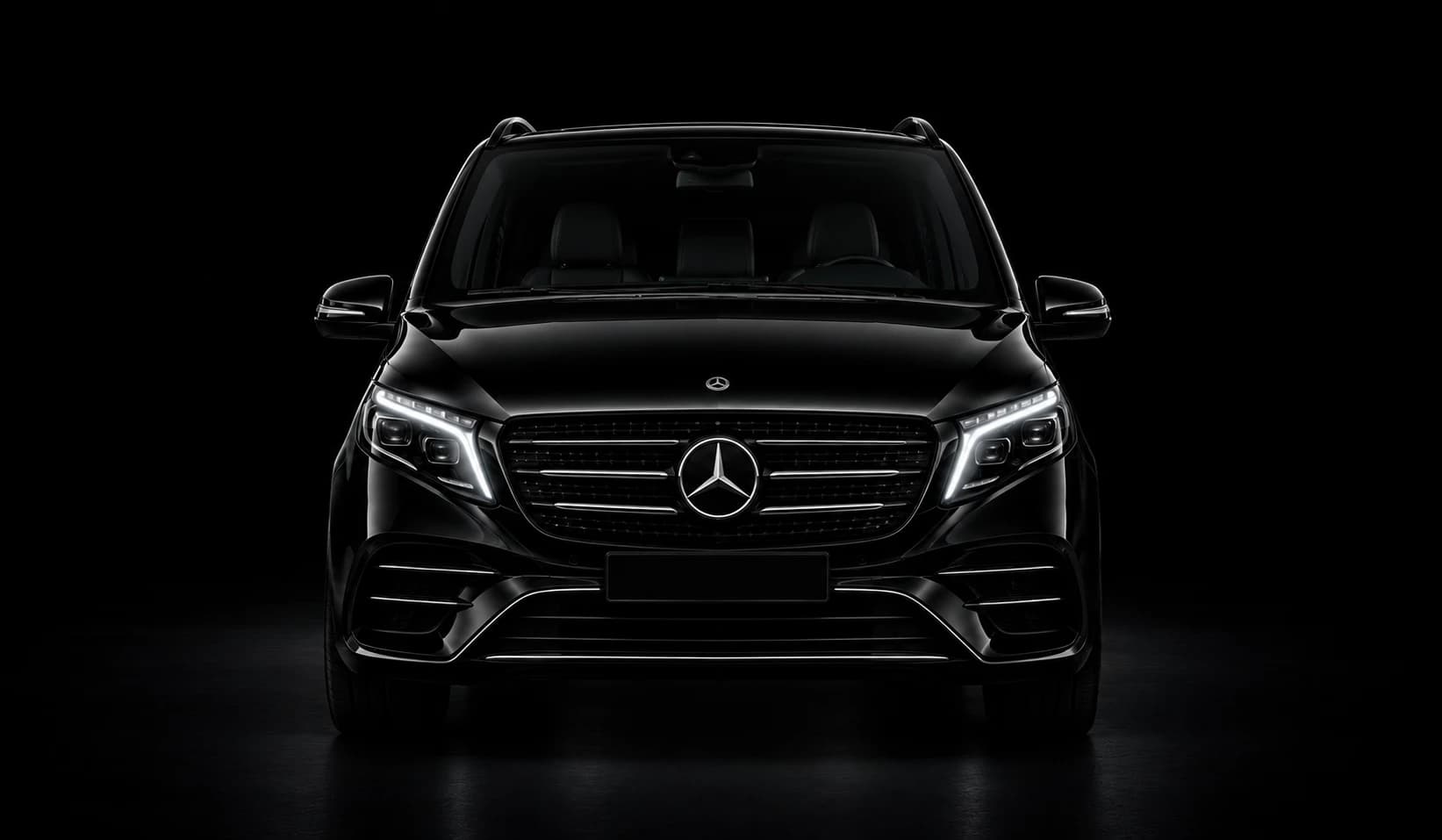 Black Mercedes-Benz V-Class people carrier, front view, headlights on, against a black studio background